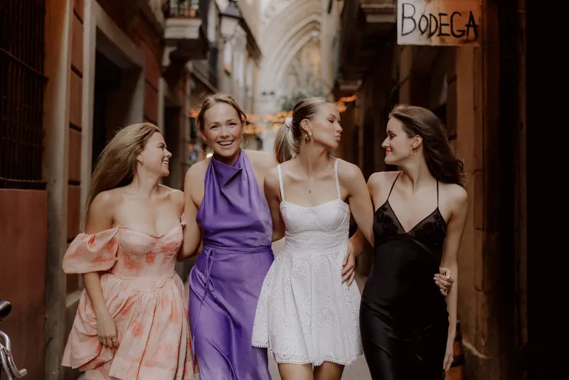 Four women in stylish dresses walk arm in arm, sharing laughter during a Vacation Portraits shoot in Barcelona’s Gothic Quart
