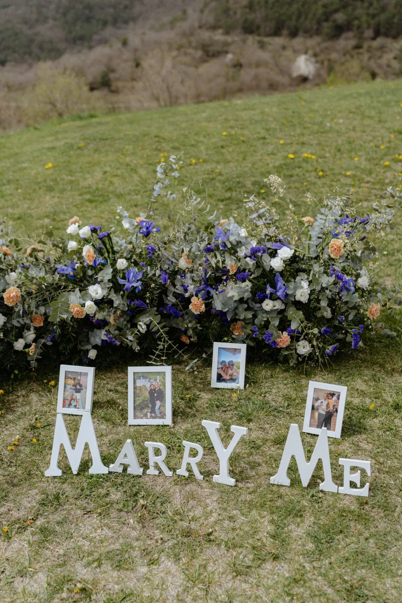 Framed couple photos and “Marry Me” letters arranged with flowers on a grassy hillside for a surprise proposal shoot.