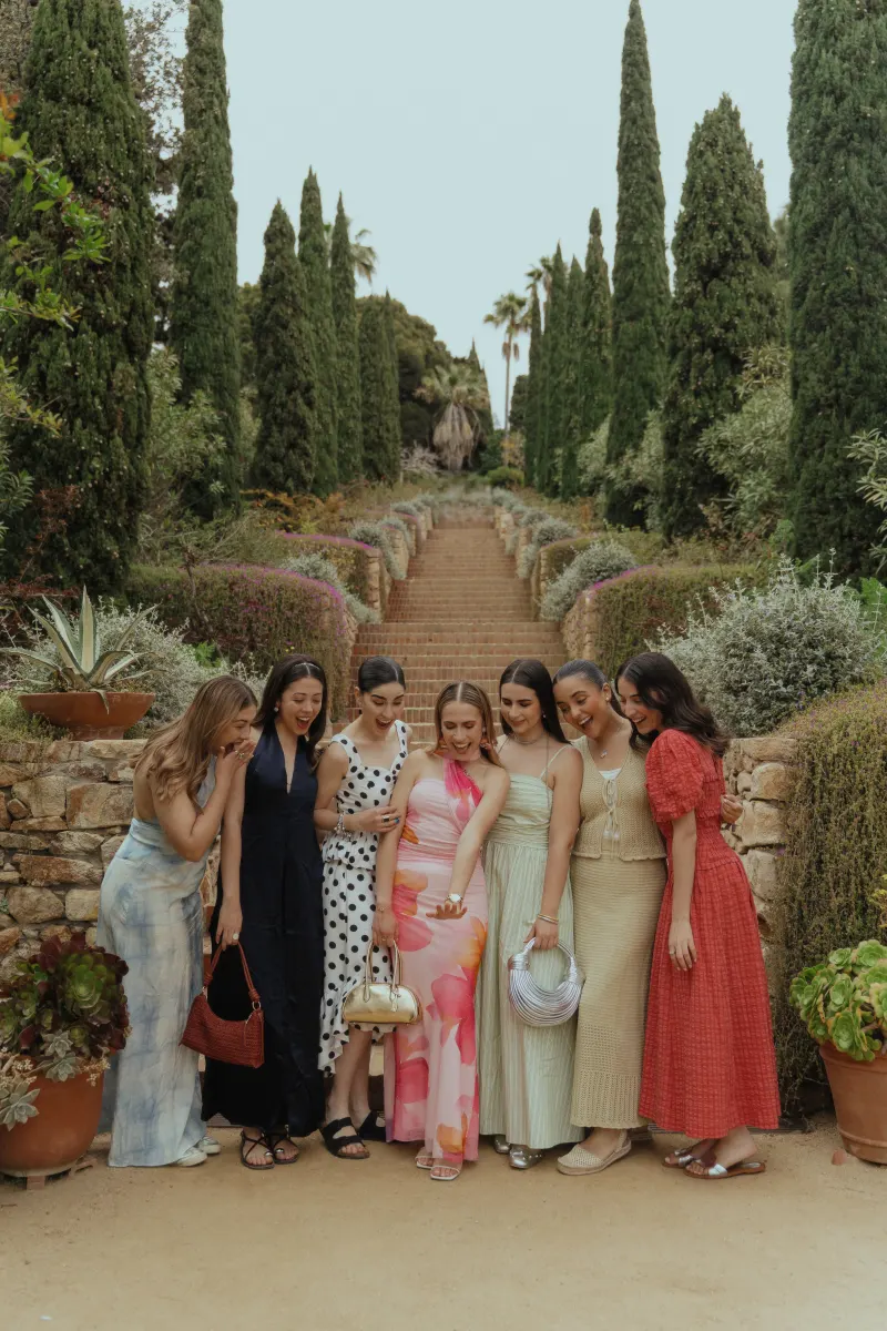 Friends gather around the bride-to-be, admiring her engagement ring during a surprise proposal photoshoot in a lush garden.