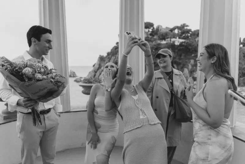 Friends laugh and celebrate with a newly engaged couple at a seaside pavilion during a surprise proposal photoshoot.