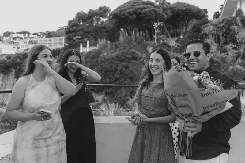 Friends laugh and wipe away happy tears as a couple celebrates their seaside surprise proposal with flowers.