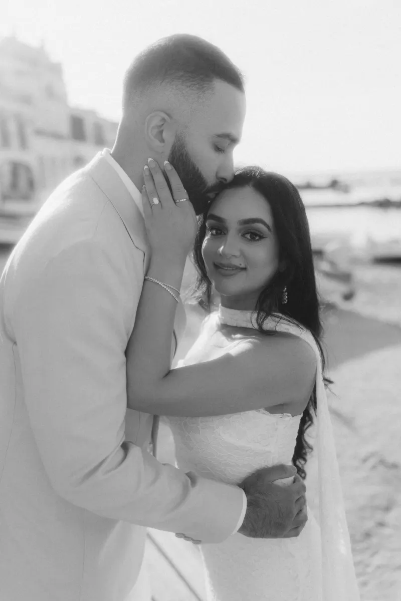 Groom-to-be kisses fiancée’s forehead during a surprise proposal photoshoot by the waterfront, capturing pure joy.