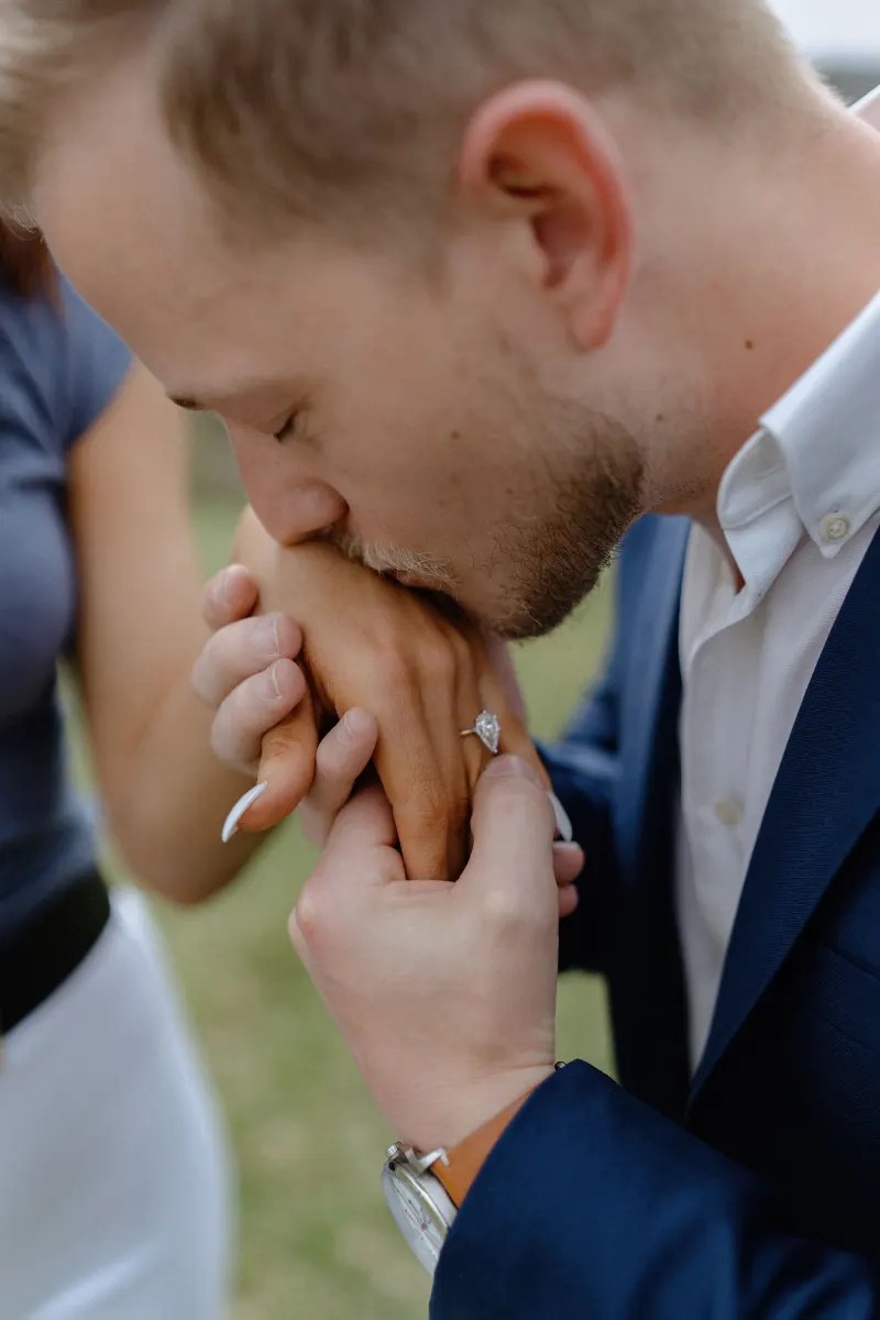 Groom-to-be kisses fiancée’s hand, highlighting her engagement ring during a surprise proposal photoshoot outdoors.