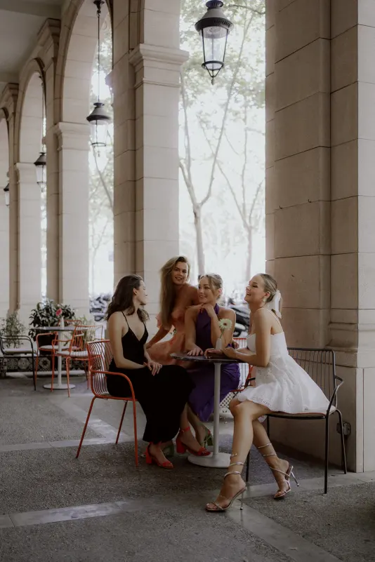Group of friends in elegant dresses share a joyful moment at a café table, Vacation Portraits photoshoot under city arches.