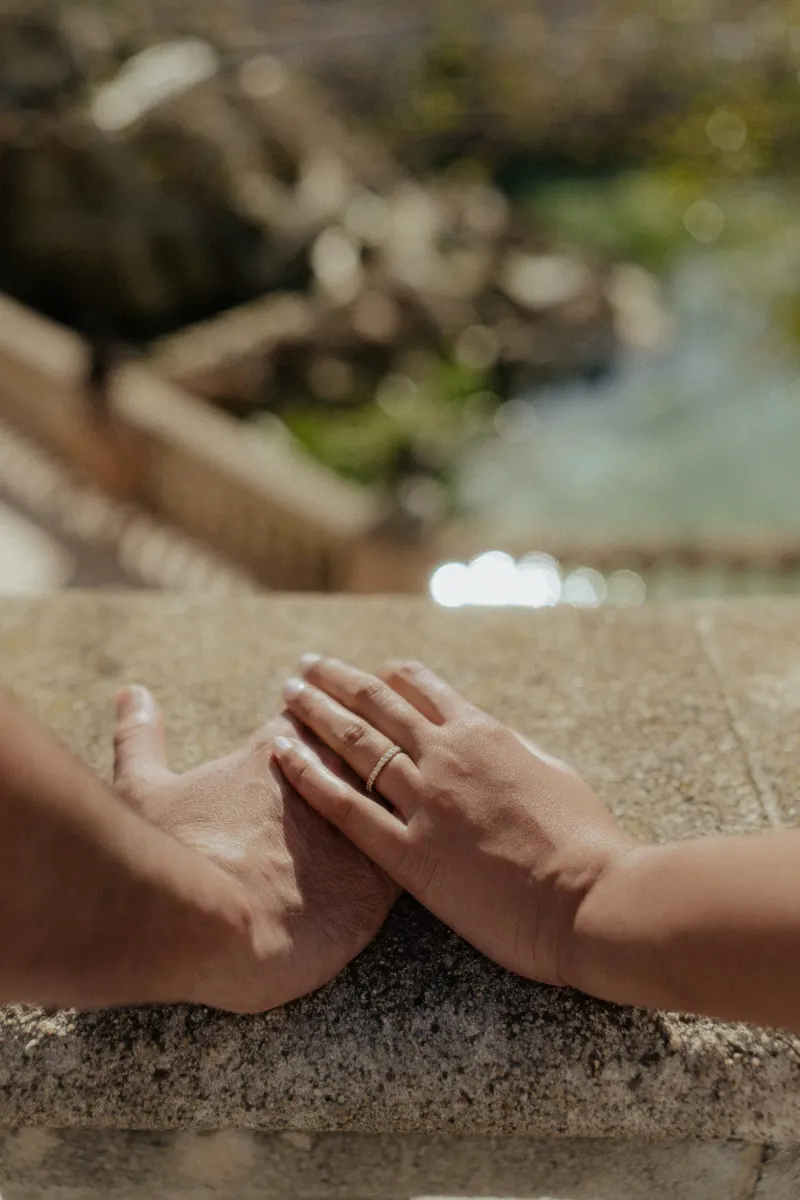 Hands gently touching on a sunlit stone ledge, engagement ring visible during a couples photoshoot outdoors.