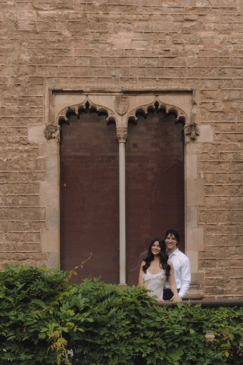 Happy couple stands together by a historic stone window, smiling during a couples & engagements photoshoot.