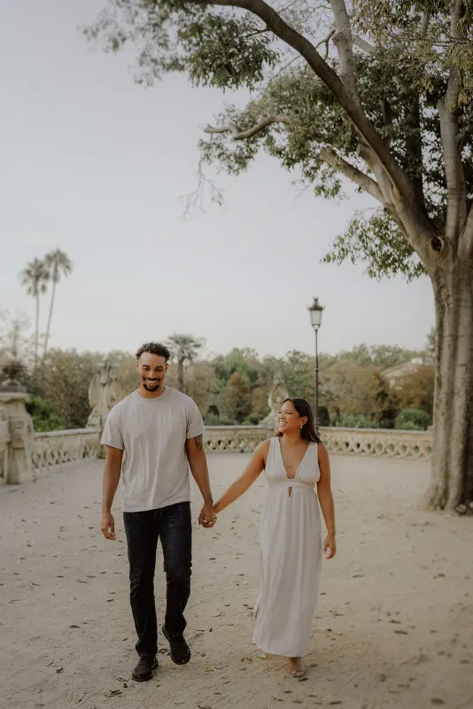Happy couple walks hand in hand through a sunlit park, sharing a candid moment during their engagement photoshoot.