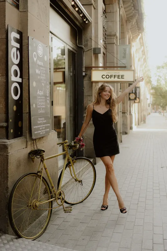 Happy woman in a black dress poses with a yellow bike outside a café during a Vacation Portraits photoshoot in Europe.