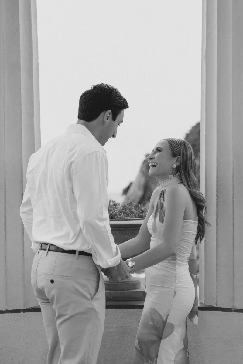 Joyful couple holding hands and laughing during a surprise proposal photoshoot at an elegant seaside pavilion.