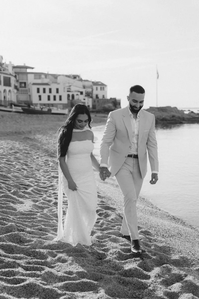 Joyful couple holding hands on a sandy beach after a surprise proposal photoshoot, with coastal village in the background.