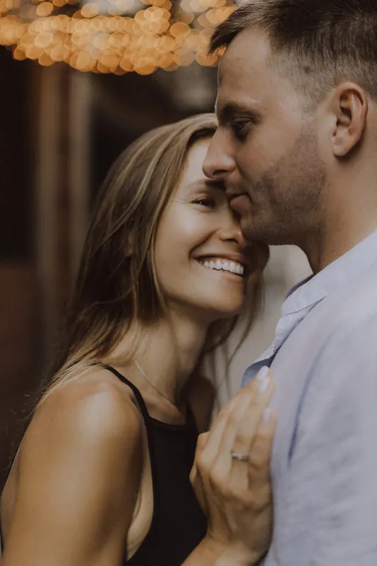 Joyful couple shares a close, loving moment under warm string lights during a couples & engagements photoshoot.