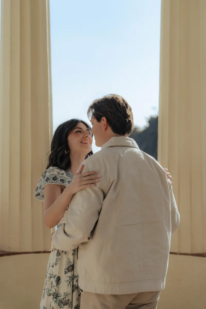 Joyful couple shares a loving embrace under sunlit columns during a surprise proposal photoshoot by the sea.