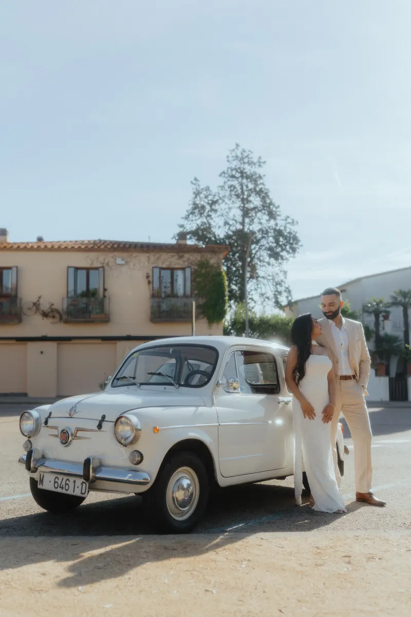 Joyful couple stands beside a classic car in a sunlit plaza, celebrating their surprise proposal photoshoot.