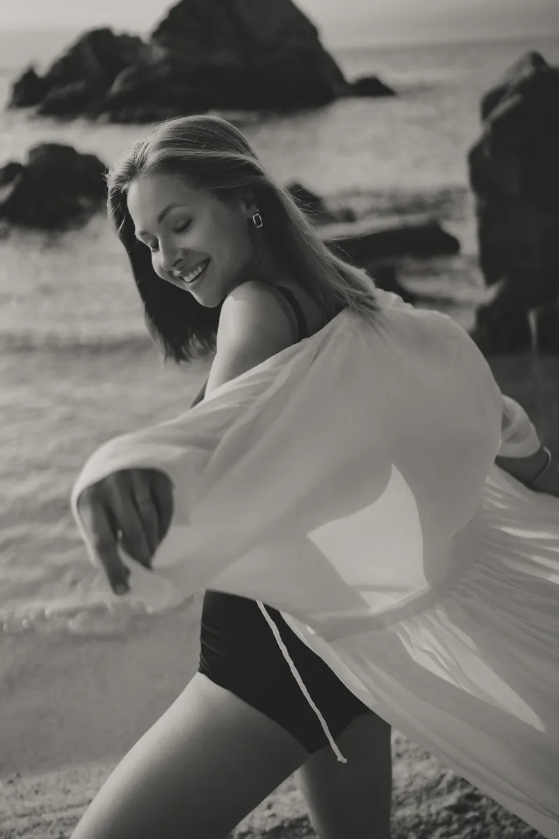 Joyful woman in swimwear and cover-up spins by the shore, captured during a Vacation Portraits photoshoot.