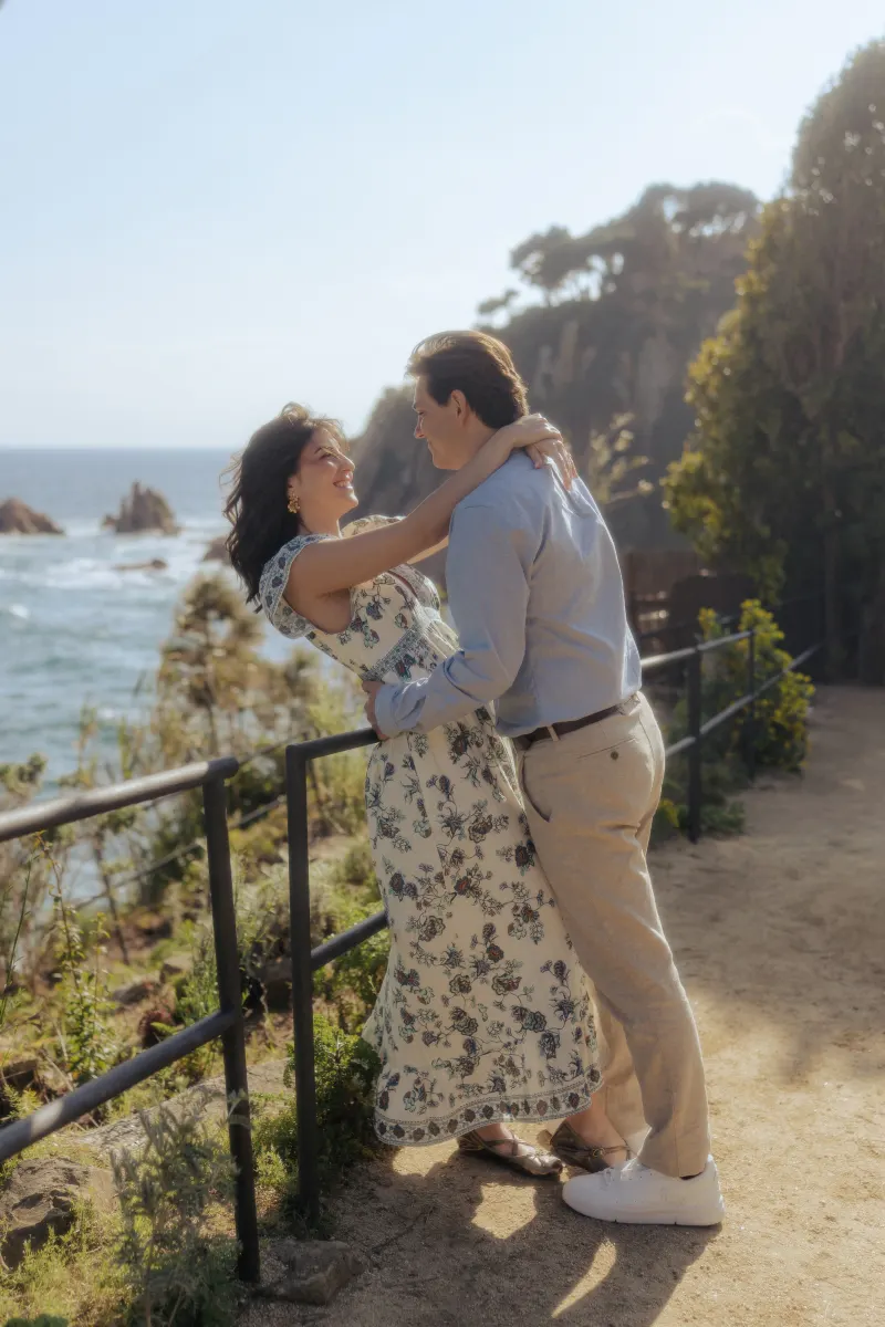 Laughing couple embraces on a coastal path, captured during a surprise proposal photoshoot at the seaside.