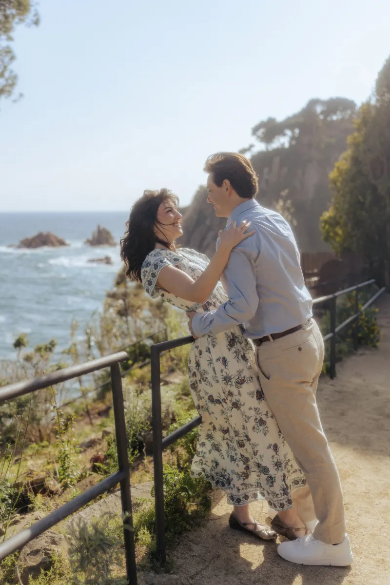 Laughing couple embraces on a coastal path, captured during a surprise proposal photoshoot by the sea.