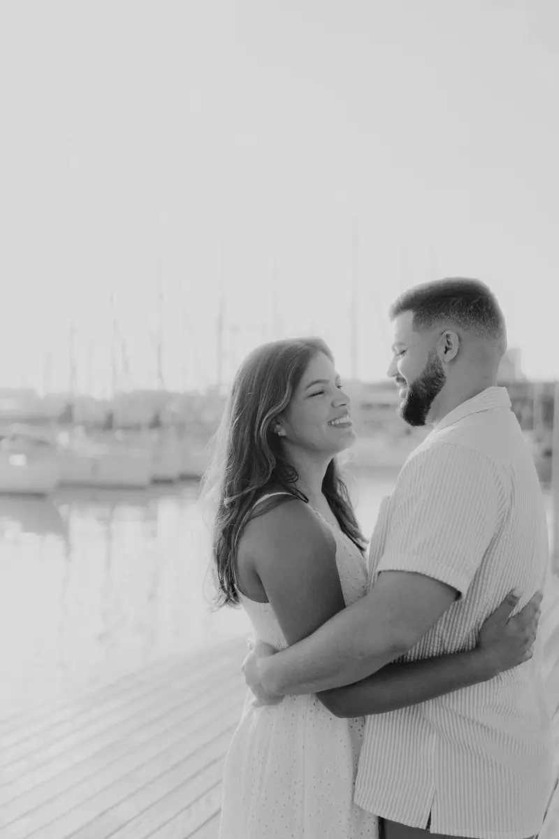 Laughing couple embraces on a sunlit marina dock during a couples and engagements photoshoot.