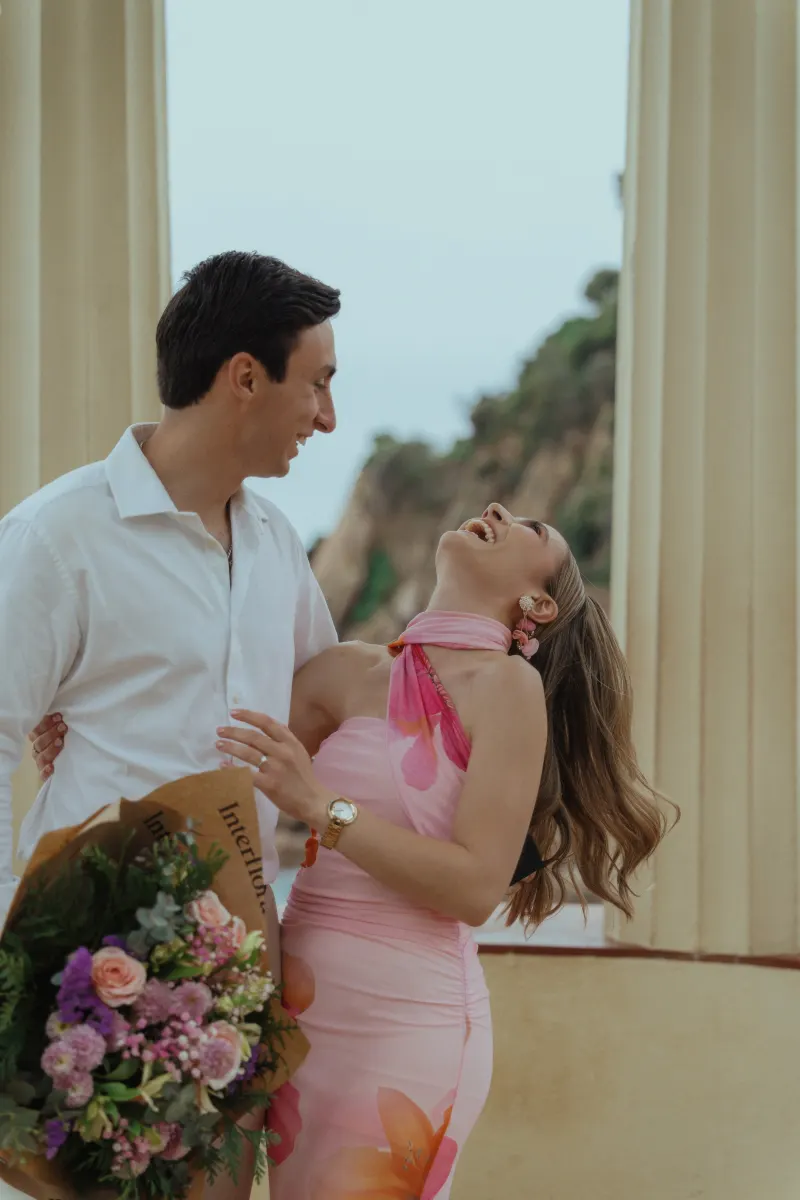 Laughing couple embraces with a bouquet at a seaside pavilion during a surprise proposal photoshoot.