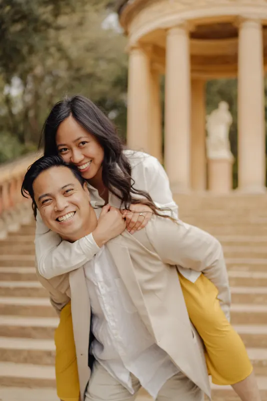 Laughing couple enjoys a playful piggyback ride on stone steps during a couples & engagements photoshoot in the park.