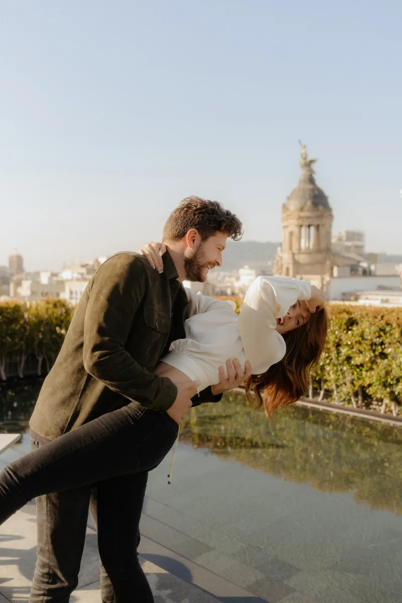 Laughing couple shares a playful dip during an engagement photoshoot on a sunny rooftop in the city.