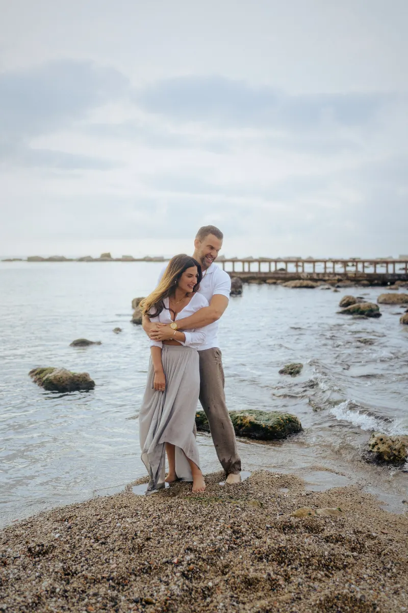 Laughing couple sharing a warm embrace on a pebbled beach, captured during a couples & engagements photoshoot.