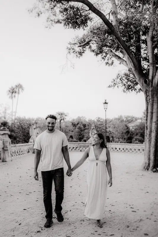 Laughing couple strolls hand in hand under leafy trees during a relaxed engagement session in a historic park setting.