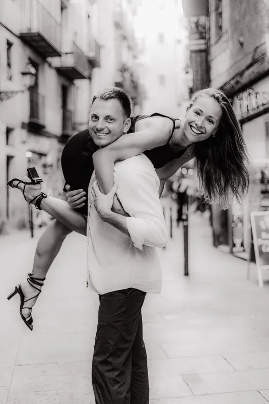 Man carries his laughing partner on his back down a city street during a playful couples & engagements photoshoot.
