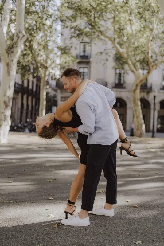 Man dips his laughing partner in a playful dance on a sunlit city street during a couples & engagements photoshoot.