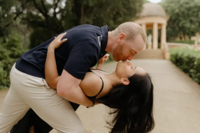 Man dips his partner for a romantic kiss on a garden path during a surprise proposal photoshoot.