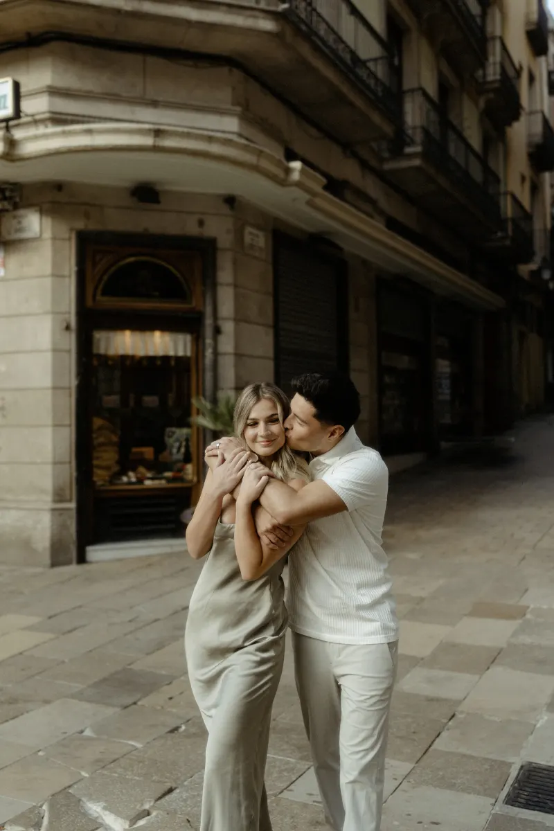 Man embraces his partner from behind and kisses her cheek on a quiet Barcelona street during a Vacation Portraits session.