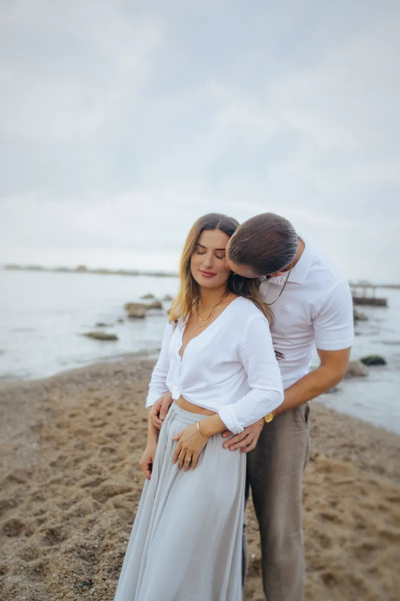 Man gently embracing his partner from behind on a peaceful beach, couples & engagements photoshoot.