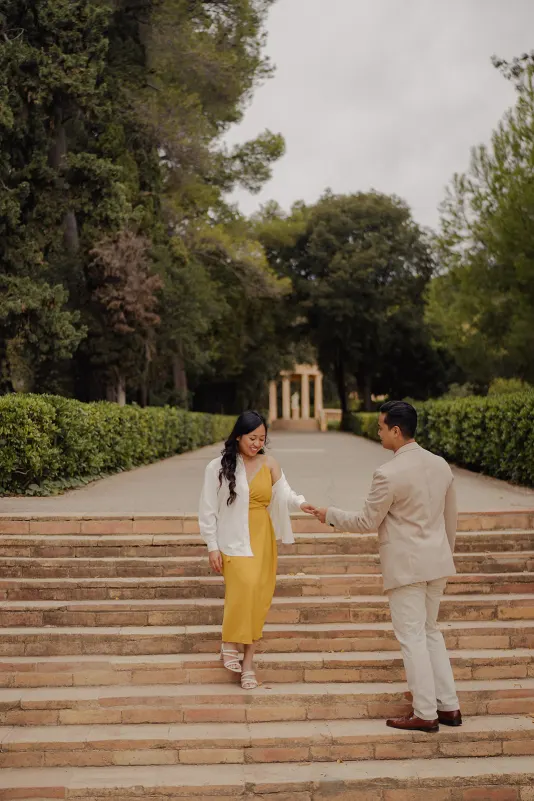 Man helps his partner down stone steps in a lush park, sharing a sweet moment during a couples & engagements photoshoot.