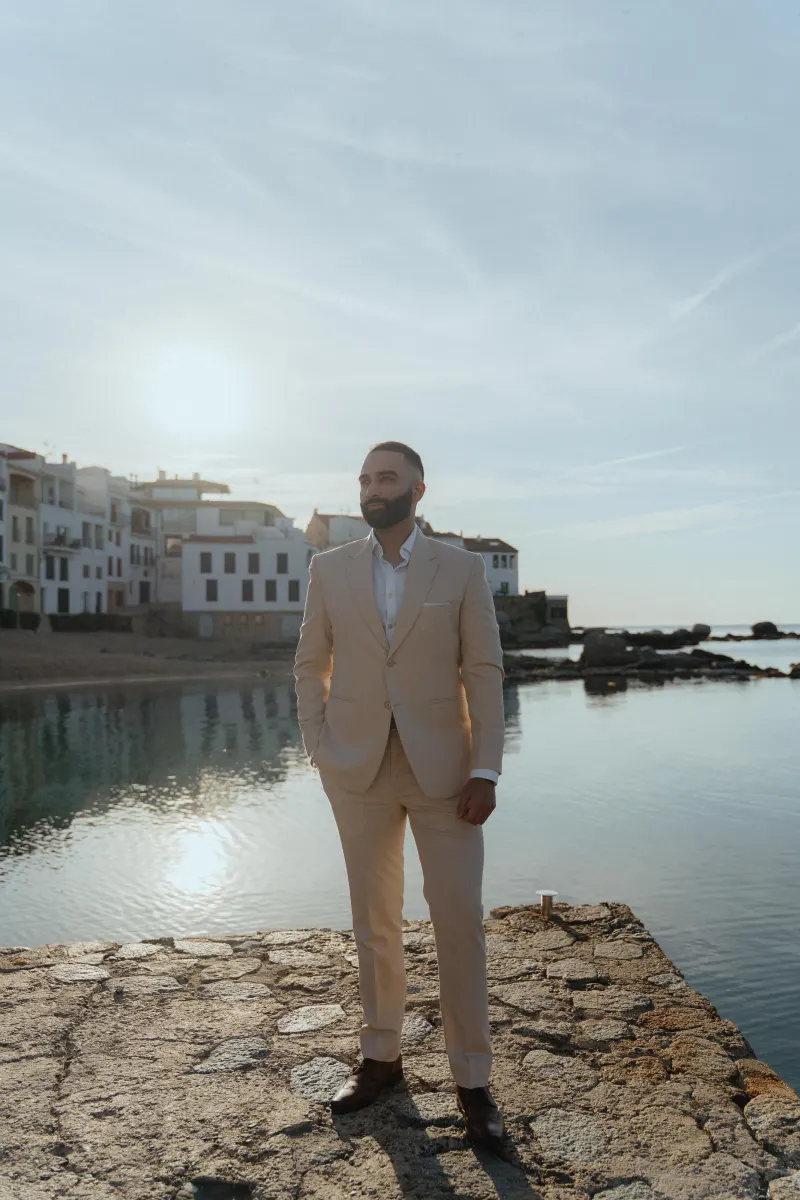 Man in a beige suit stands by the water at sunset, ready for a surprise proposal photoshoot by the coast.