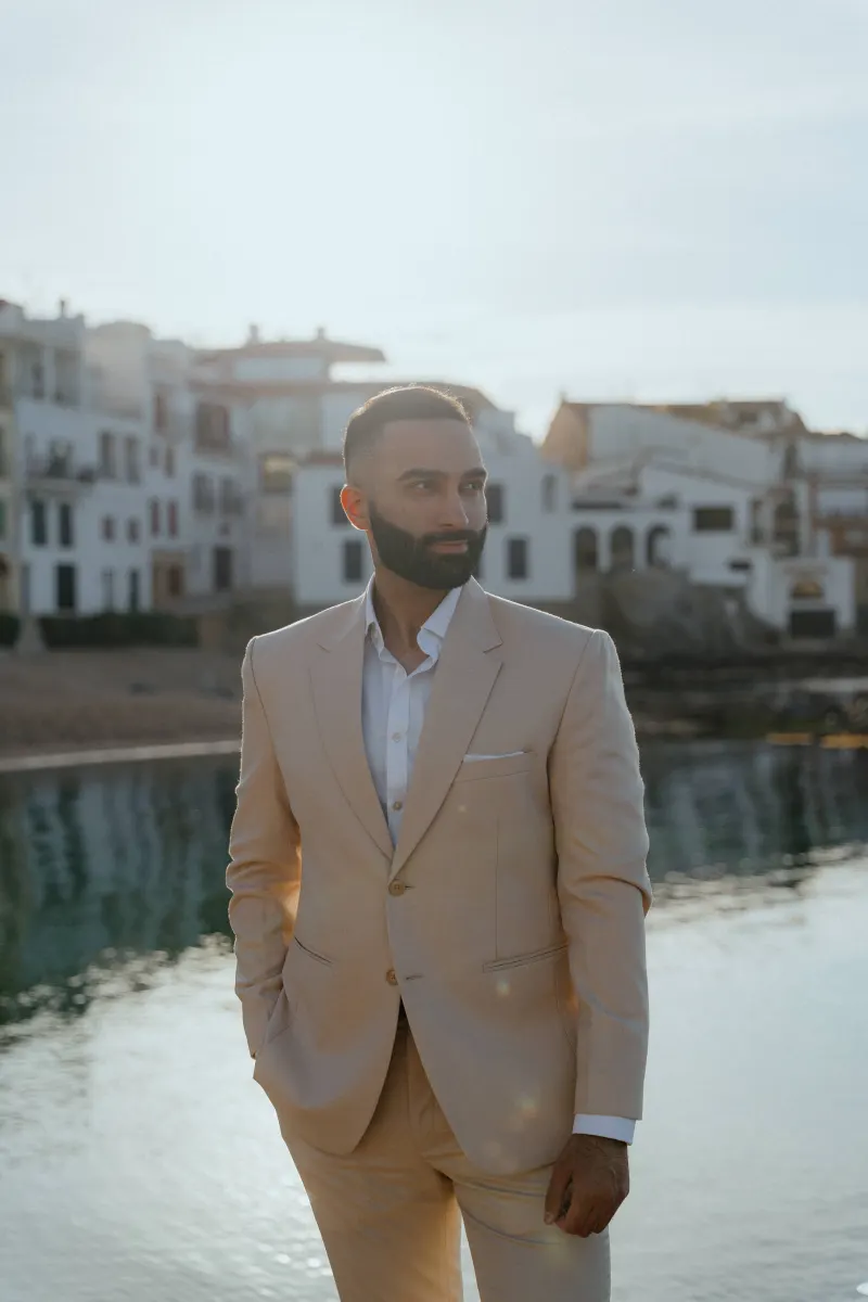 Man in a beige suit stands by the waterfront at sunset, ready for a surprise proposal photoshoot in a coastal village.