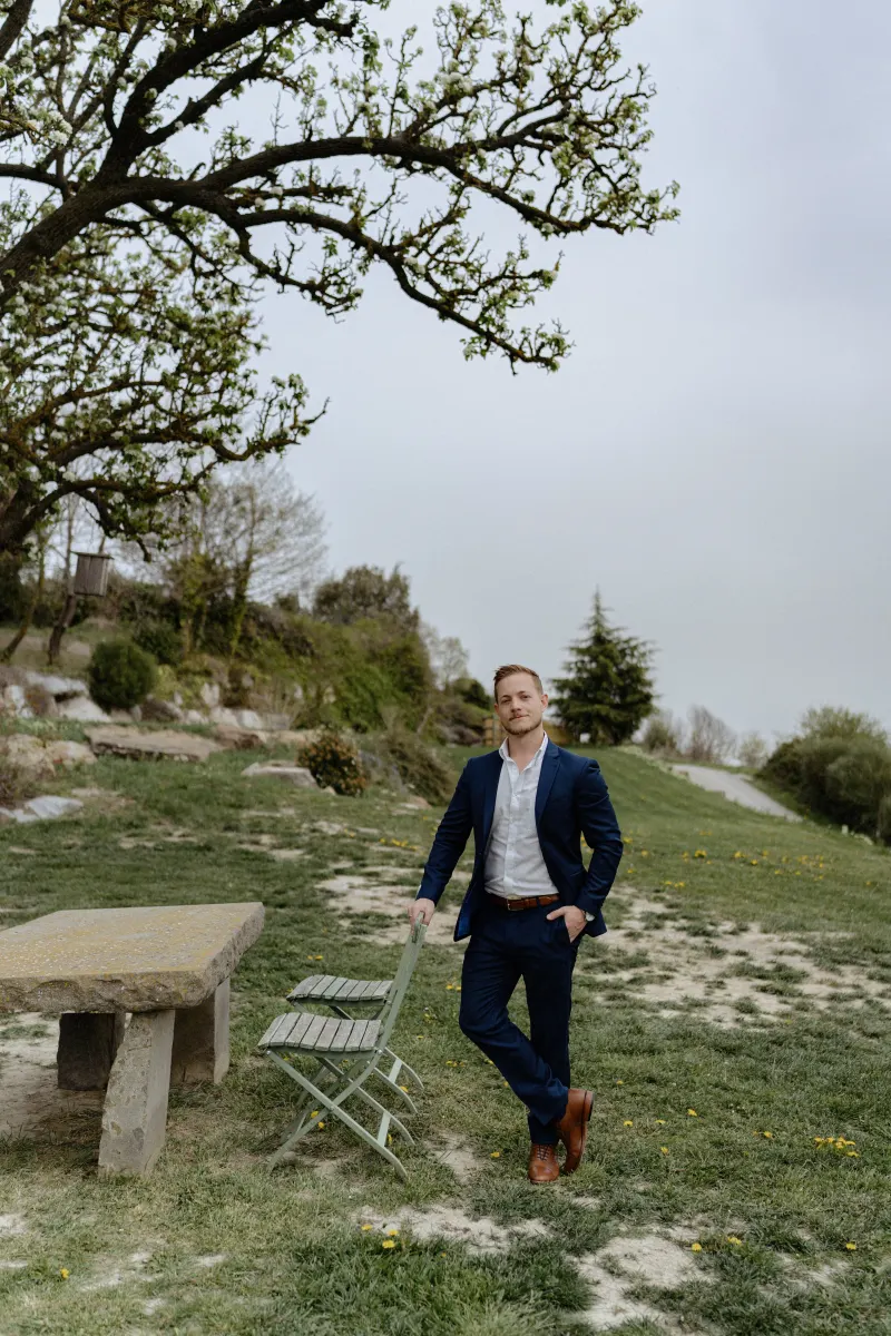 Man in a navy suit waits by a rustic bench under a blooming tree, ready for a countryside surprise proposal photoshoot.