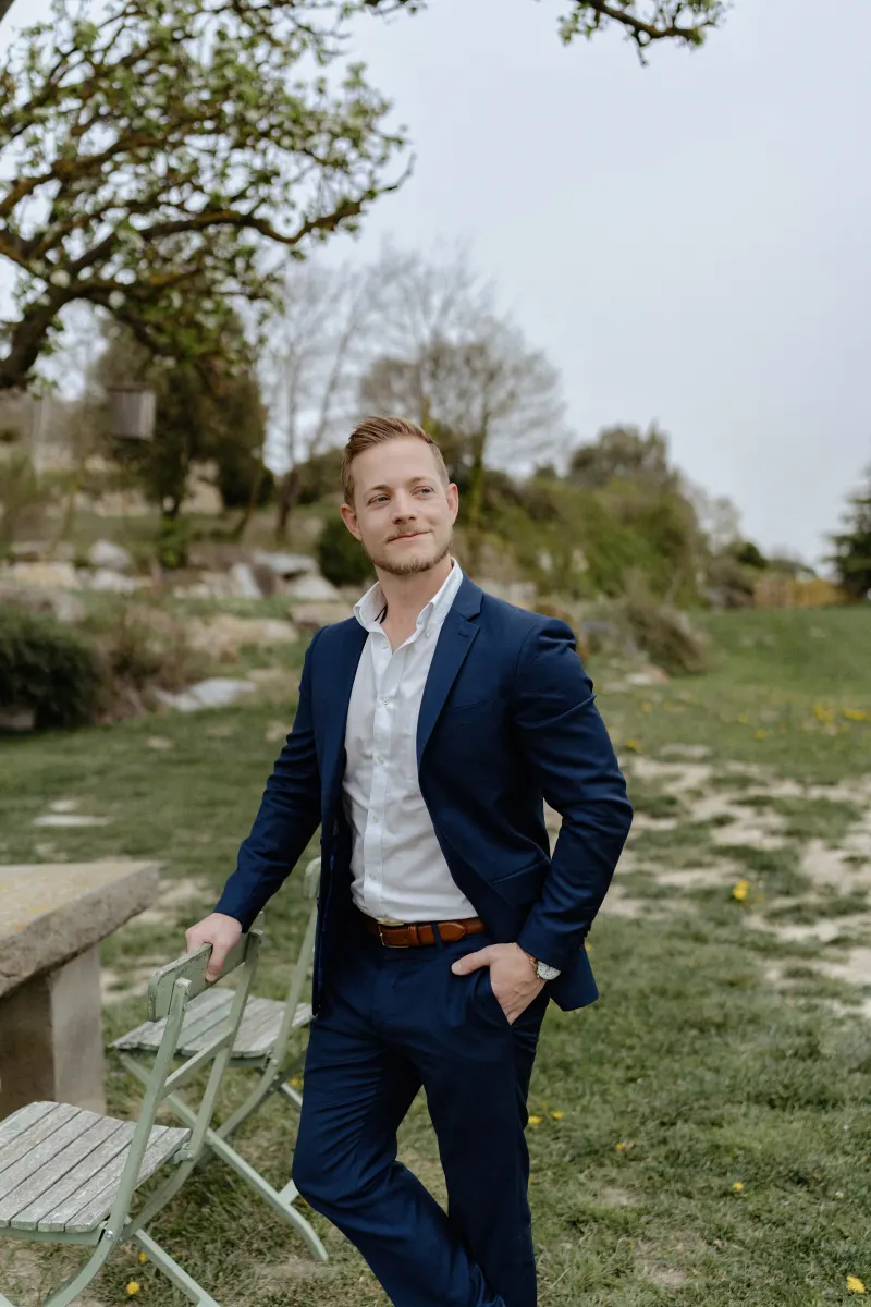 Man in a navy suit waits with a hopeful smile in a countryside garden before a surprise proposal photoshoot.