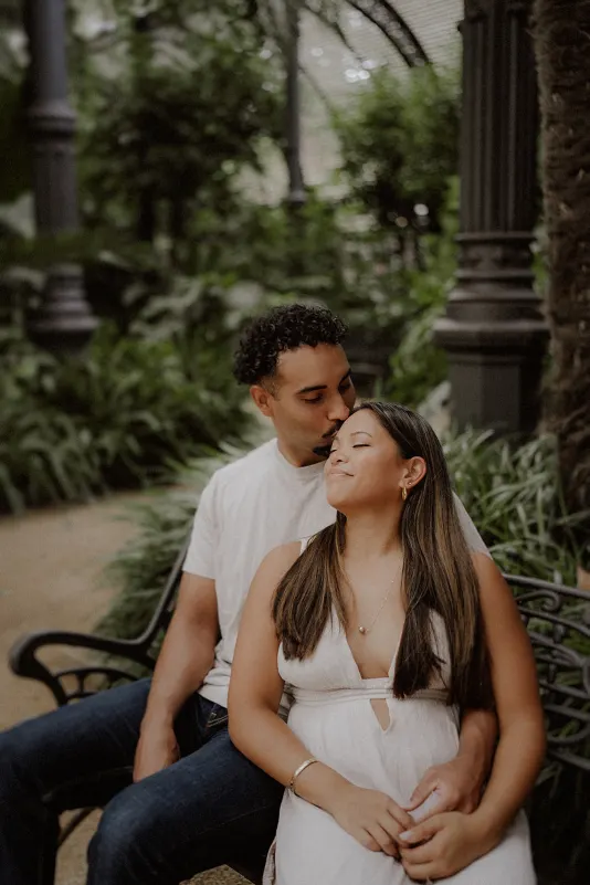 Man kisses woman’s forehead as they sit close on a garden bench during a couples & engagements photoshoot.