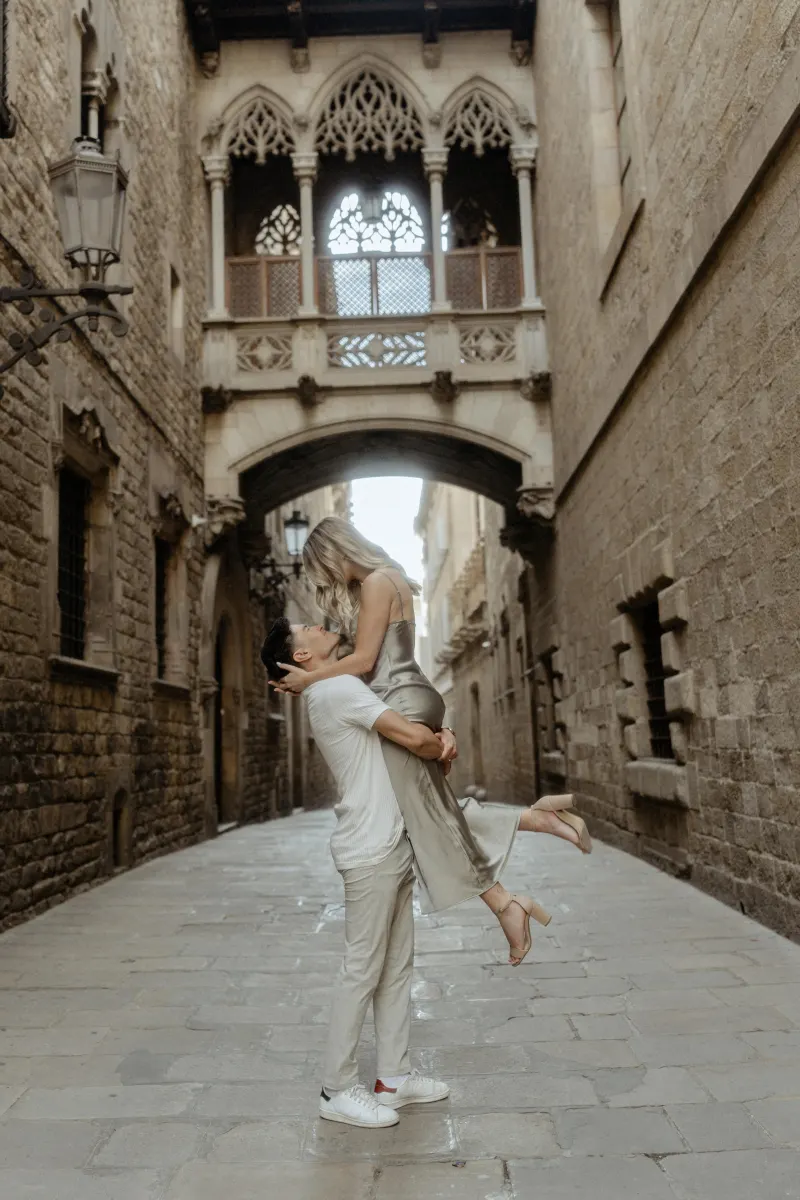Man lifts his partner in a joyful embrace beneath Barcelona’s Gothic bridge during a Vacation Portraits photoshoot.