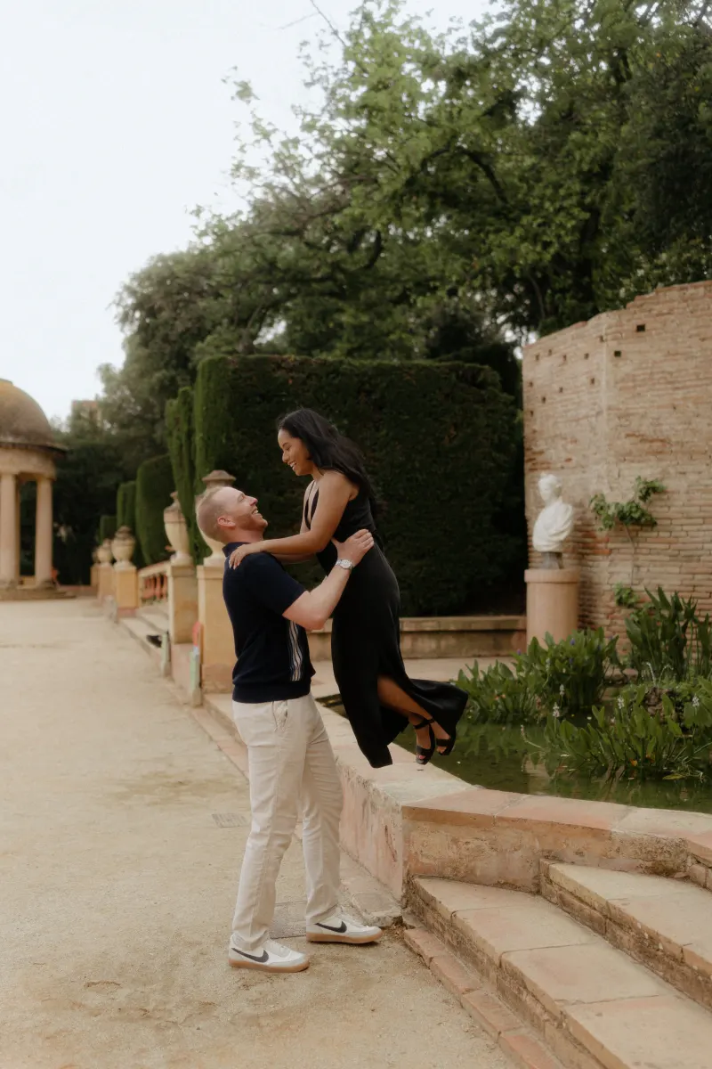 Man lifts his partner in a joyful embrace beside a garden pond during a surprise proposal photoshoot.