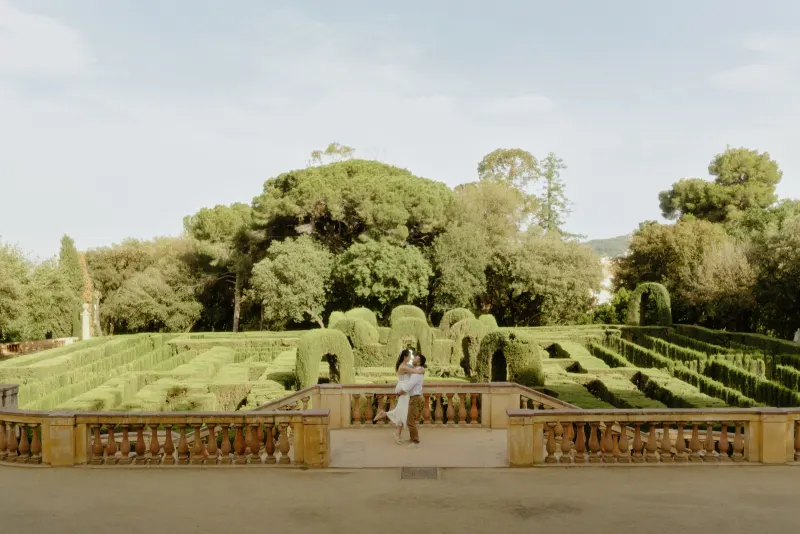 Man lifts his partner in a joyful embrace on a sunlit terrace overlooking a lush garden maze during a couples photoshoot.
