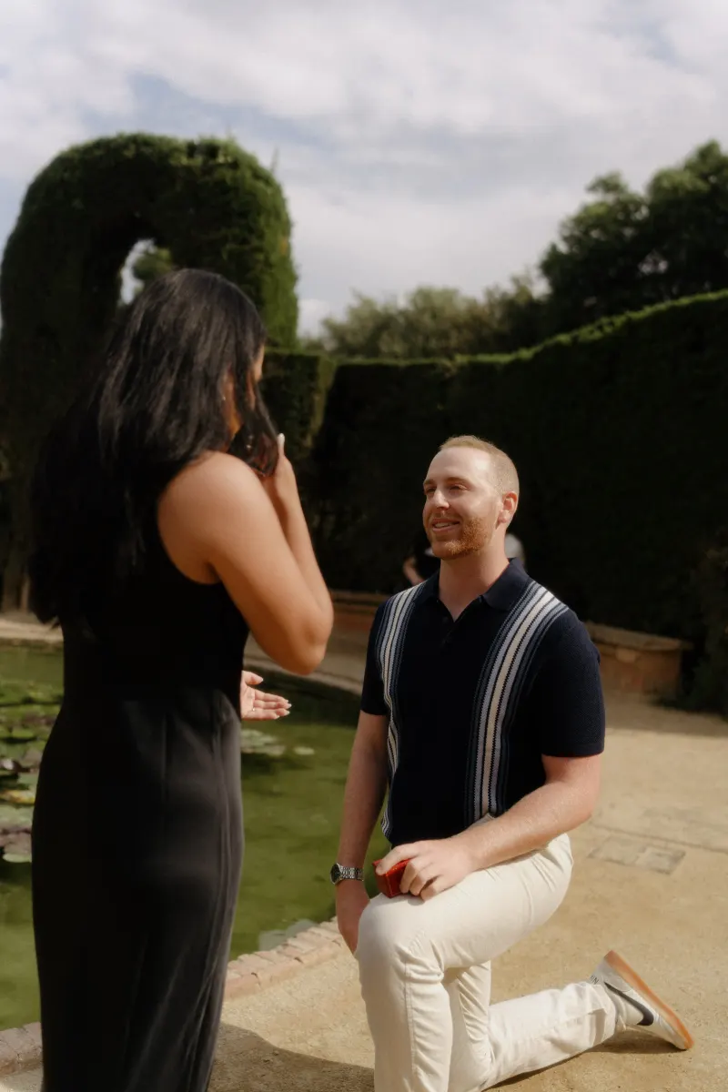 Man on one knee proposes to his surprised partner by a pond, captured during a surprise proposal photoshoot outdoors.