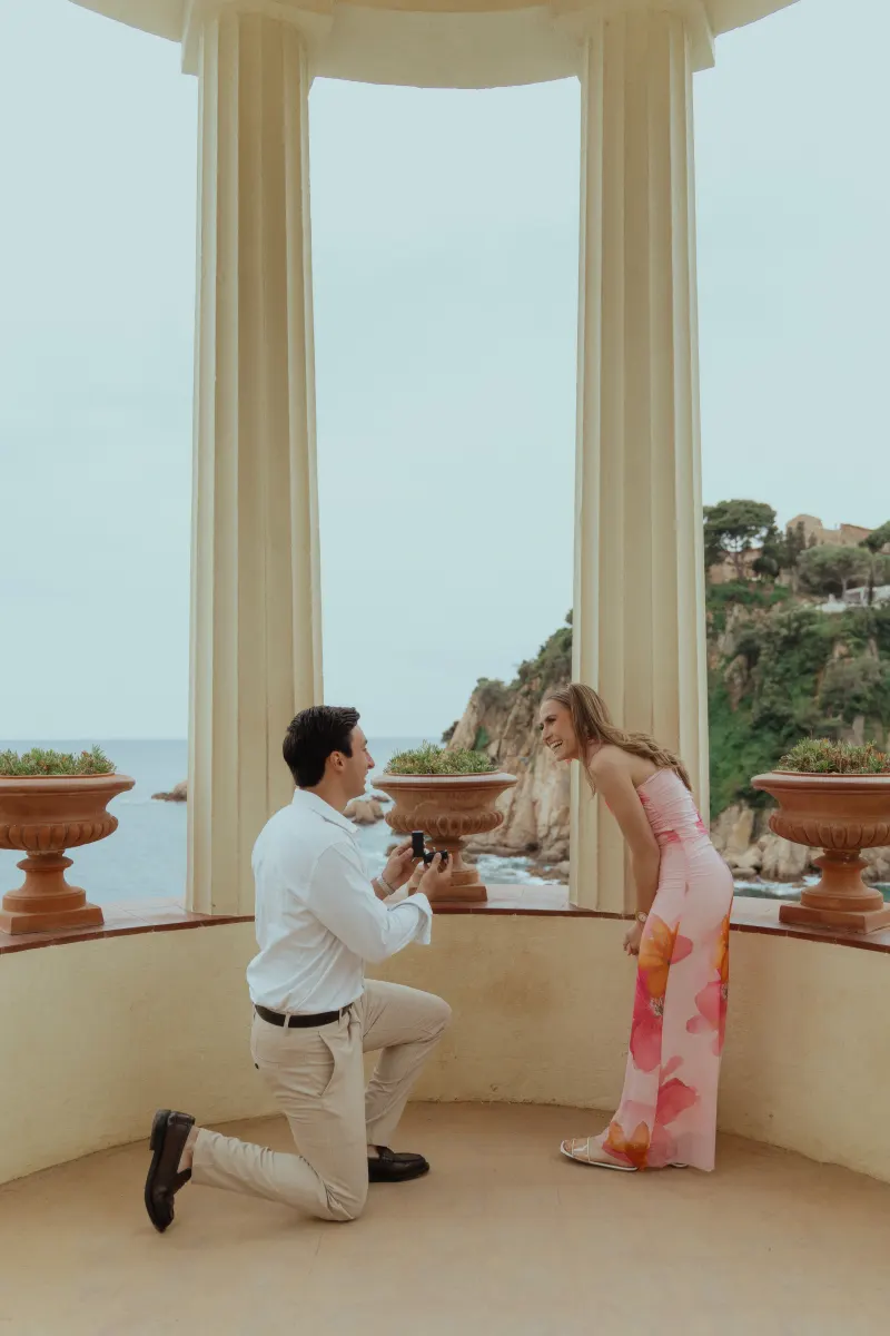 Man on one knee proposing to his smiling partner in a seaside pavilion, captured during a surprise proposal photoshoot.