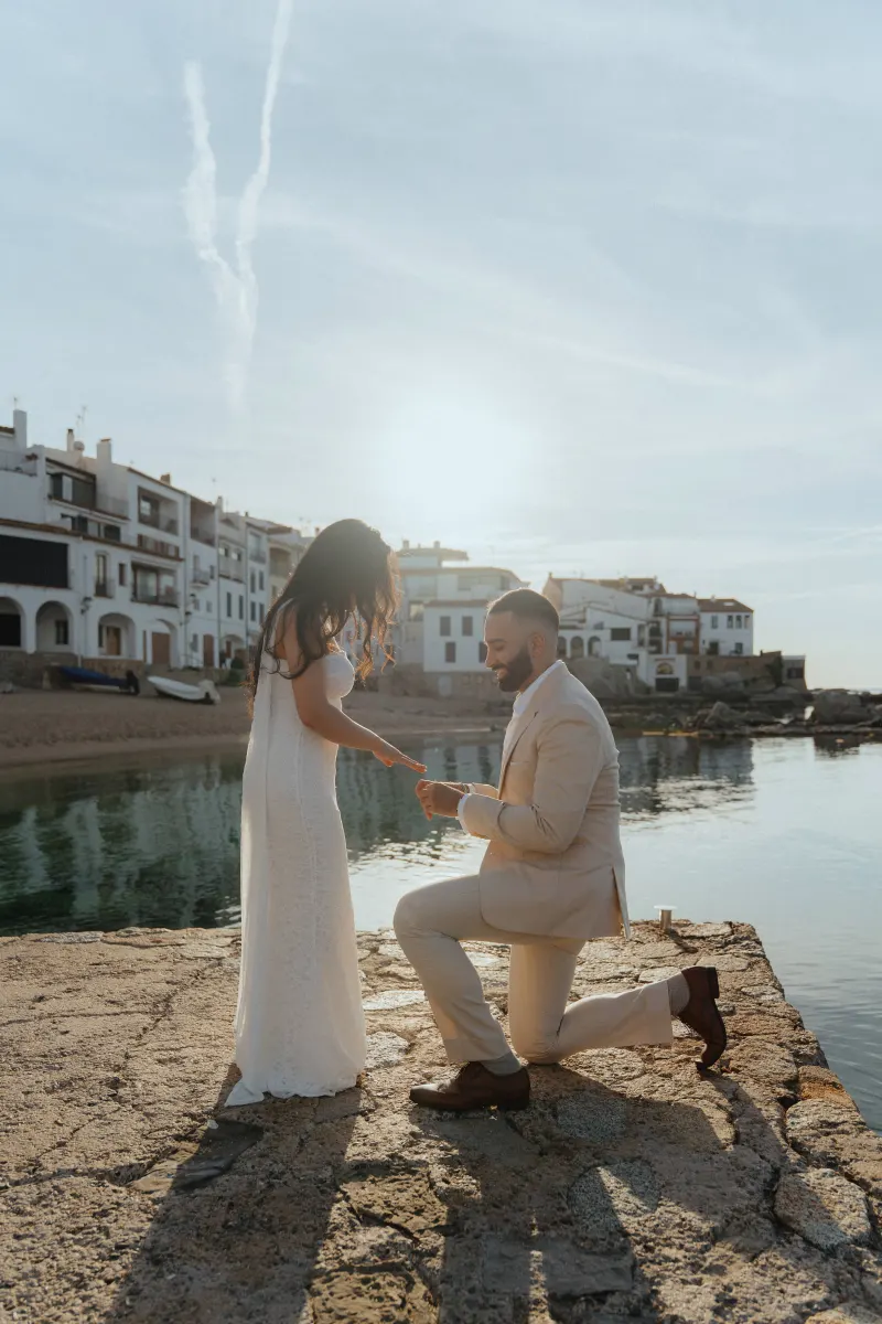 Man proposes on a sunlit pier by the water, capturing a joyful surprise proposal photoshoot moment.