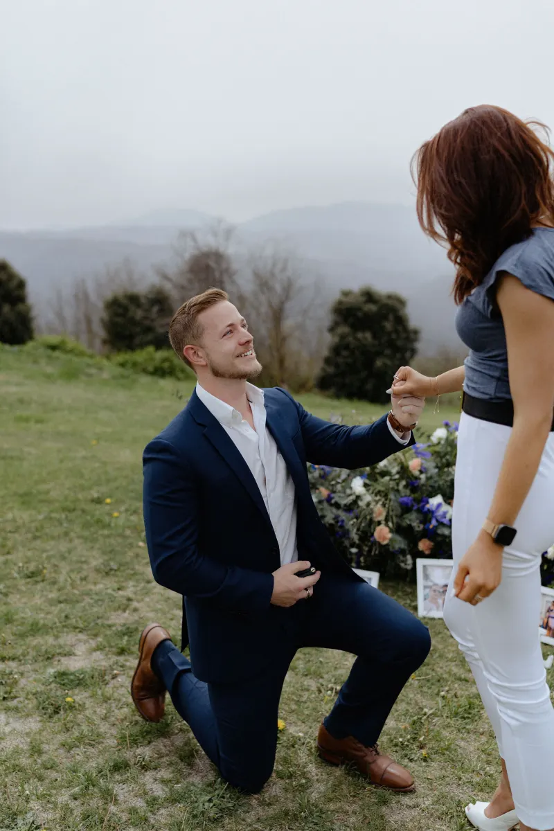Man proposes on one knee, holding his partner’s hand on a scenic hillside during a surprise proposal photoshoot.