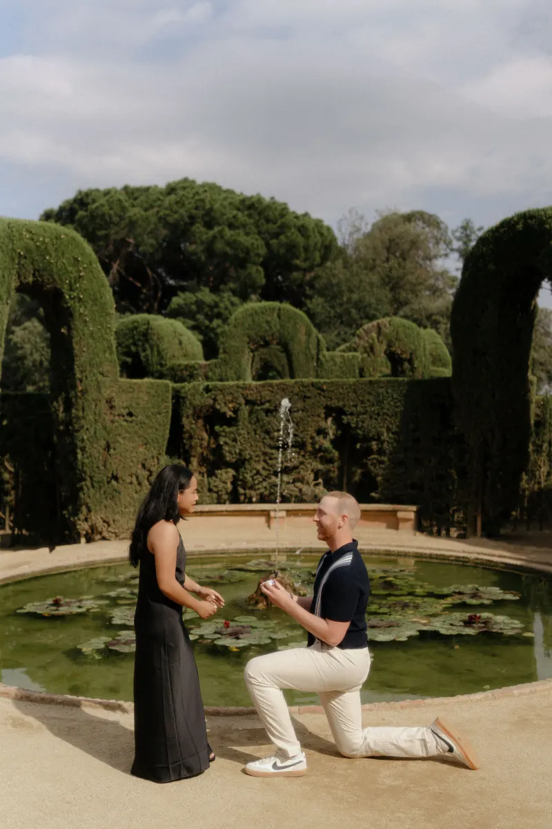 Man proposes on one knee to his partner beside a lily pond, captured during a surprise proposal photoshoot in a garden.