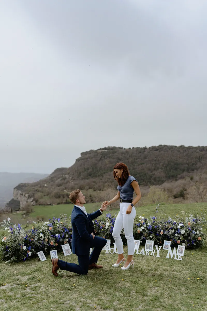 Man proposes on one knee to his partner on a flower-lined hillside, capturing a joyful moment during a surprise proposal phot