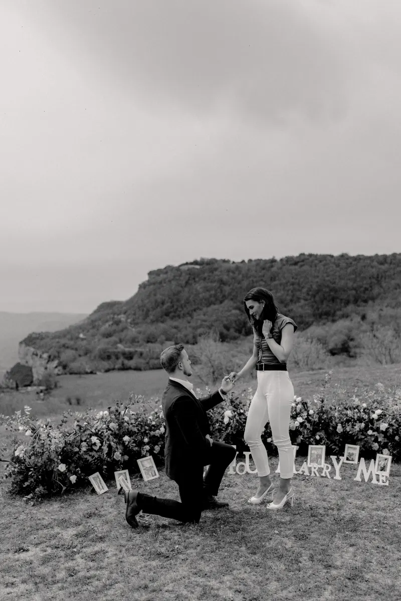 Man proposes on one knee to his partner on a flower-lined hillside, capturing joy during a surprise proposal photoshoot.