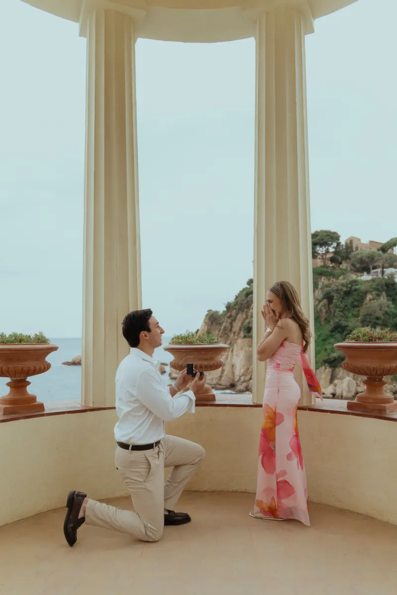 Man proposes on one knee to his surprised partner in a romantic seaside pavilion, captured during a proposal photoshoot.