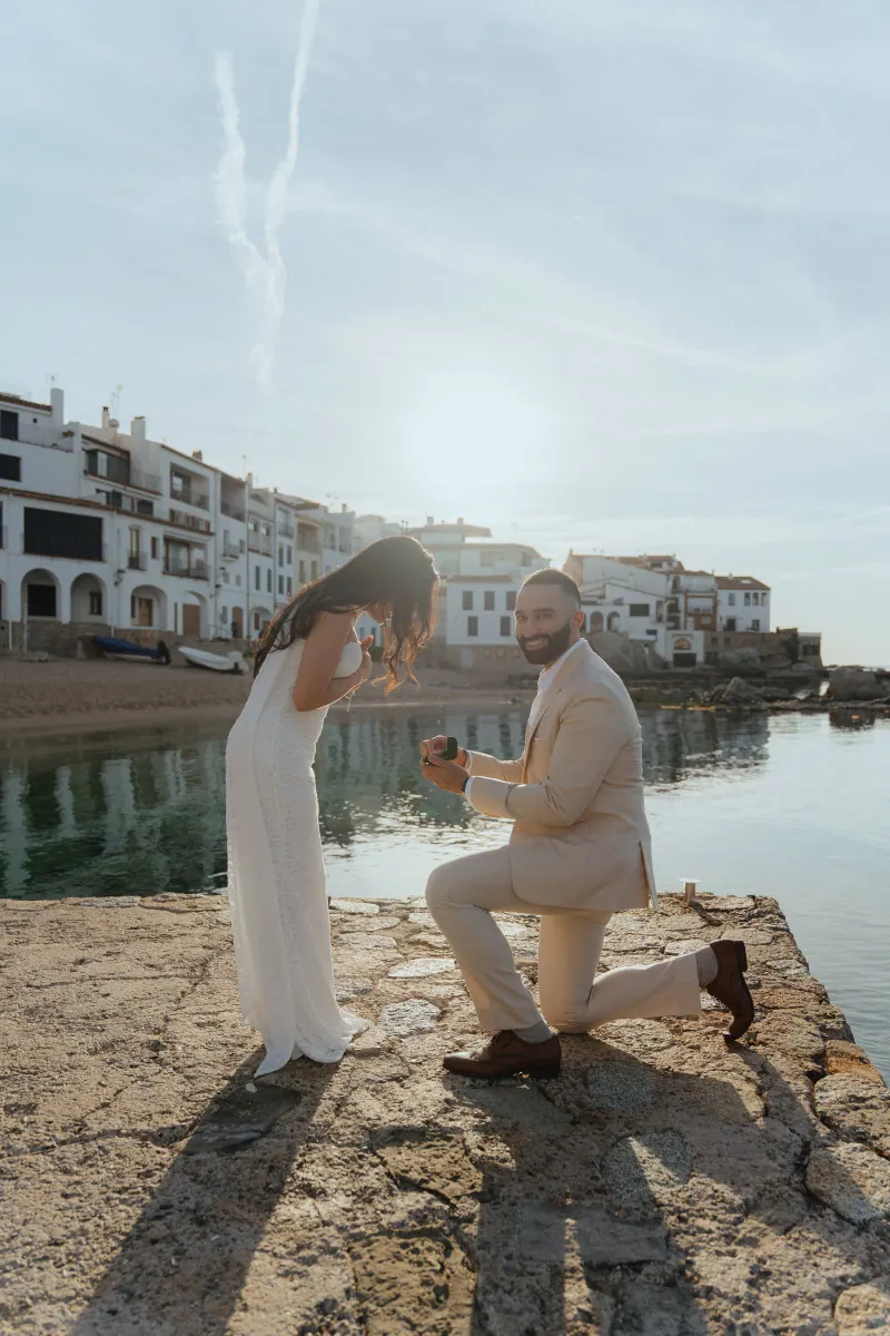 Man proposes to his partner by the water at sunset, capturing a joyful surprise proposal photoshoot in a coastal village.