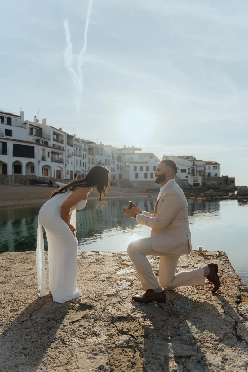 Man proposing on a sunny waterfront, capturing a joyful surprise proposal photoshoot in a charming coastal village.